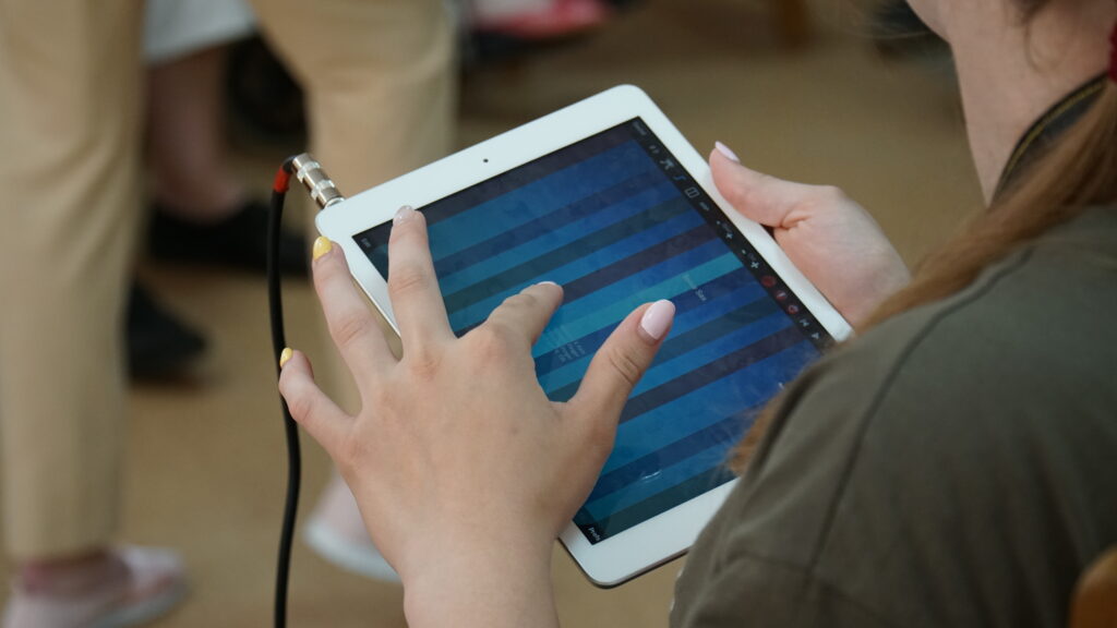Två händer håller i iPad och ena handen spelar på en skärm.